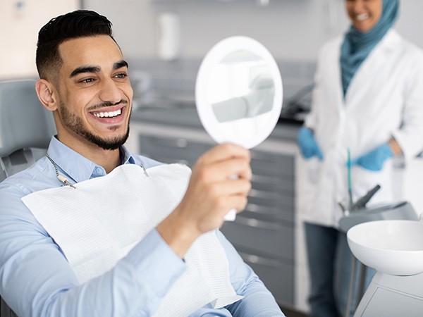 Man smiling into handheld mirror with dentist blurry in background